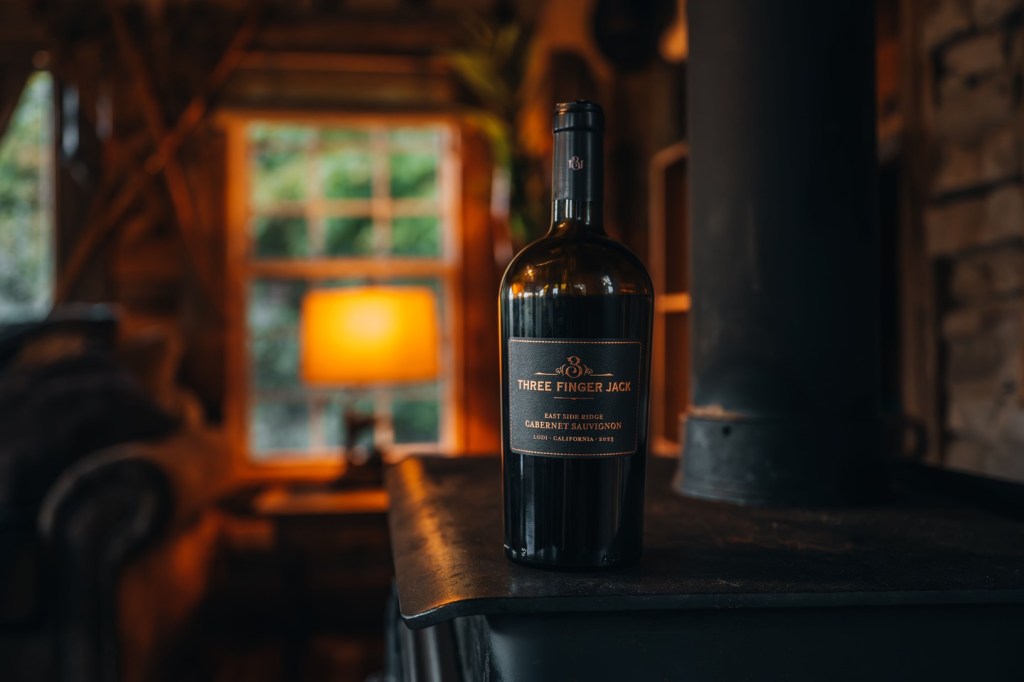 A bottle of Three Finger Jack Cabernet Sauvignon pictured on a wood burning stove inside a cozy cabin